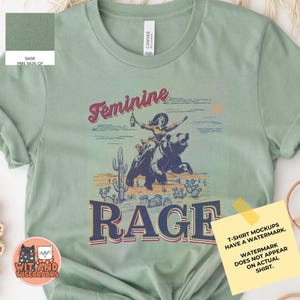 May include: Sage green t-shirt with a vintage-style graphic of a woman riding a bear with the text "Feminine Rage".