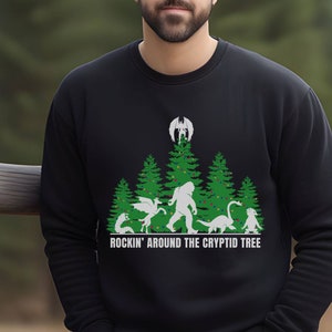 Puede incluir: Sudadera negra con un dise&ntilde;o gr&aacute;fico que presenta un &aacute;rbol de Navidad con varios cr&iacute;ptidos, incluyendo a Bigfoot y un drag&oacute;n. El texto "ROCKIN' AROUND THE CRYPTID TREE" est&aacute; impreso debajo. La sudadera es de material suave.
