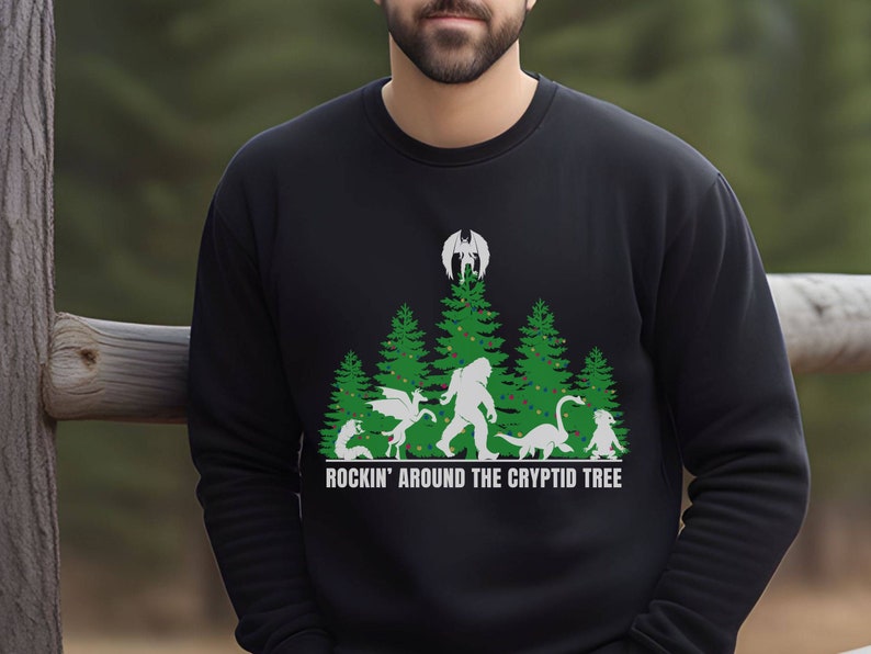 Puede incluir: Sudadera negra con un dise&ntilde;o gr&aacute;fico que presenta un &aacute;rbol de Navidad con varios cr&iacute;ptidos, incluyendo a Bigfoot y un drag&oacute;n. El texto "ROCKIN' AROUND THE CRYPTID TREE" est&aacute; impreso debajo. La sudadera es de material suave.