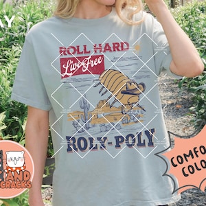 May include: A light blue t-shirt with a graphic of a roly-poly bug wearing a cowboy hat in a desert scene. The text "Roll Hard Live Free Roly-Poly" is printed on the shirt. The shirt is available in comfort colors.