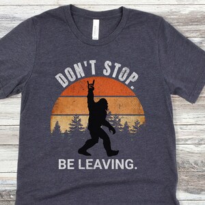 K&ouml;nnte beinhalten: Ein dunkel-helles graues T-Shirt mit einer Vintage-Grafik eines Bigfoot-Silhouetten, das seine Hand in einer Rock'n'Roll-Geste hebt. Die Grafik ist vor einem Sonnenuntergangs-Hintergrund mit B&auml;umen und dem Text "Don't Stop. Be Leaving." platziert.