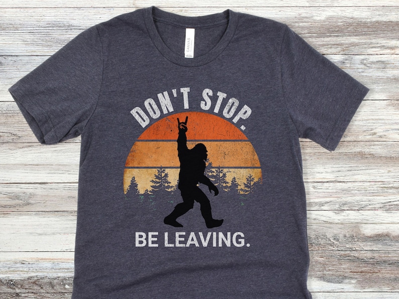 K&ouml;nnte beinhalten: Ein dunkel-helles graues T-Shirt mit einer Vintage-Grafik eines Bigfoot-Silhouetten, das seine Hand in einer Rock'n'Roll-Geste hebt. Die Grafik ist vor einem Sonnenuntergangs-Hintergrund mit B&auml;umen und dem Text "Don't Stop. Be Leaving." platziert.