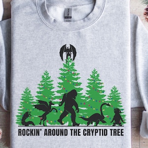 Könnte beinhalten: Hellgraues Sweatshirt mit einem grünen Aufdruck von Weihnachtsbäumen und Kryptiden. Der Text "ROCKIN' AROUND THE CRYPTID TREE" ist unten in Weiß gedruckt. Die Grafik zeigt einen Drachen, Bigfoot und das Ungeheuer von Loch Ness.