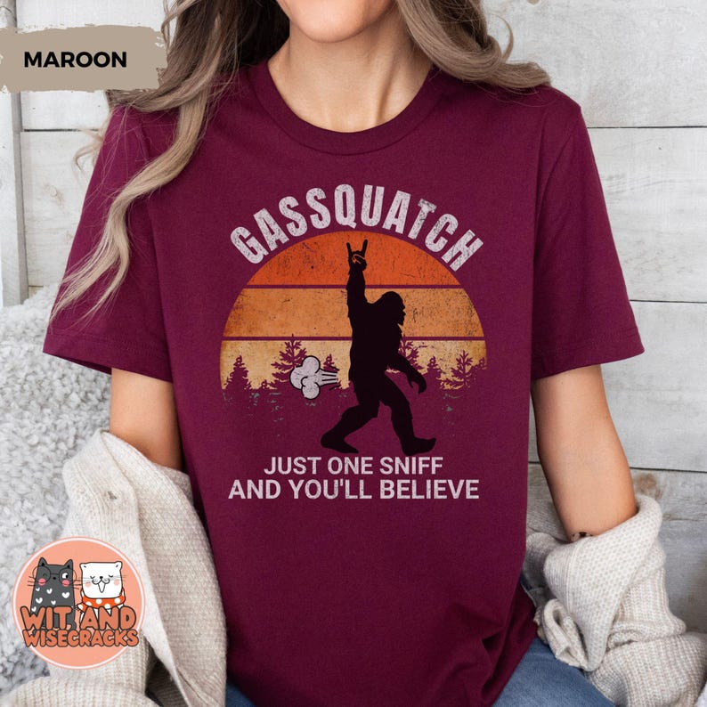 May include: Maroon t-shirt with a graphic featuring a silhouette of Bigfoot with the text "Gassquatch" above and "Just One Sniff And You'll Believe" below. The design includes a sunset and trees.