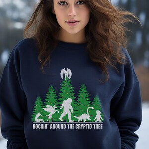 Puede incluir: Sudadera azul marino con un dise&ntilde;o de &aacute;rbol de Navidad con cr&iacute;ptidos, incluyendo un drag&oacute;n, Bigfoot y un monstruo del lago Ness. El texto "ROCKIN' AROUND THE CRYPTID TREE" est&aacute; impreso debajo.