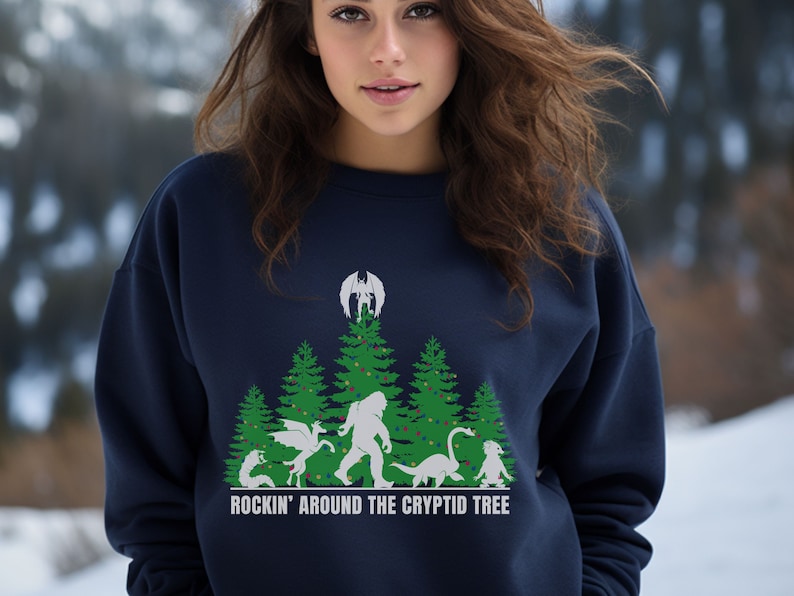 Puede incluir: Sudadera azul marino con un dise&ntilde;o de &aacute;rbol de Navidad con cr&iacute;ptidos, incluyendo un drag&oacute;n, Bigfoot y un monstruo del lago Ness. El texto "ROCKIN' AROUND THE CRYPTID TREE" est&aacute; impreso debajo.