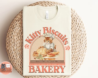May include: A cream-colored t-shirt with a vintage-style graphic of a cat baking biscuits. The graphic features a cat wearing a chef's hat and apron, kneading dough in a bowl. The text reads "Kitty Biscuits" in a pink and orange font, with the words "PAWS-ITIVELY DELIGHTFUL TREATS" and "WE KNEAD LOVE INTO EVERY BATCH" in smaller text below. The graphic is surrounded by a pink circle with a paw print on each side. The text "BAKERY" is in large, pink letters at the bottom of the graphic.