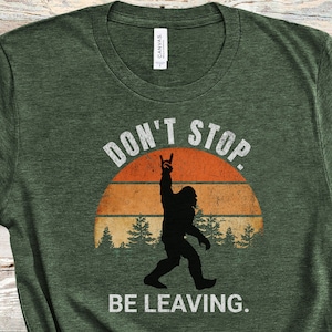 K&ouml;nnte beinhalten: Gr&uuml;nes T-Shirt mit einem Retro-Sonnenuntergang-Motiv und einer Silhouette von Bigfoot, der die Hand in die Luft hebt. Der Text "Don't Stop Be Leaving." ist auf das T-Shirt gedruckt.