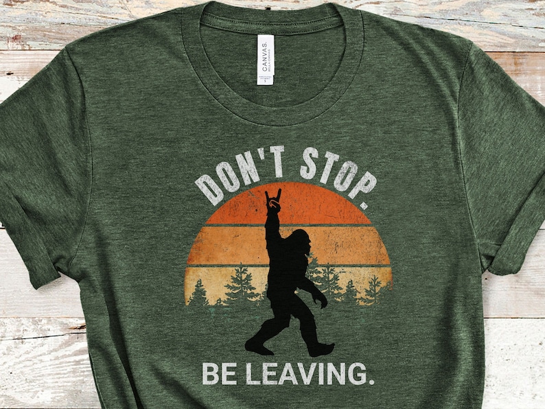 K&ouml;nnte beinhalten: Gr&uuml;nes T-Shirt mit einem Retro-Sonnenuntergang-Motiv und einer Silhouette von Bigfoot, der die Hand in die Luft hebt. Der Text "Don't Stop Be Leaving." ist auf das T-Shirt gedruckt.