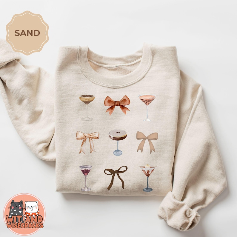 May include: A beige sweatshirt with a design of various cocktails and bows. The design is in shades of brown, orange, and white.