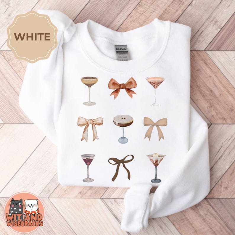 May include: White sweatshirt with a graphic design of cocktails and bows. The cocktails are illustrated in a watercolor style and the bows are in shades of brown.