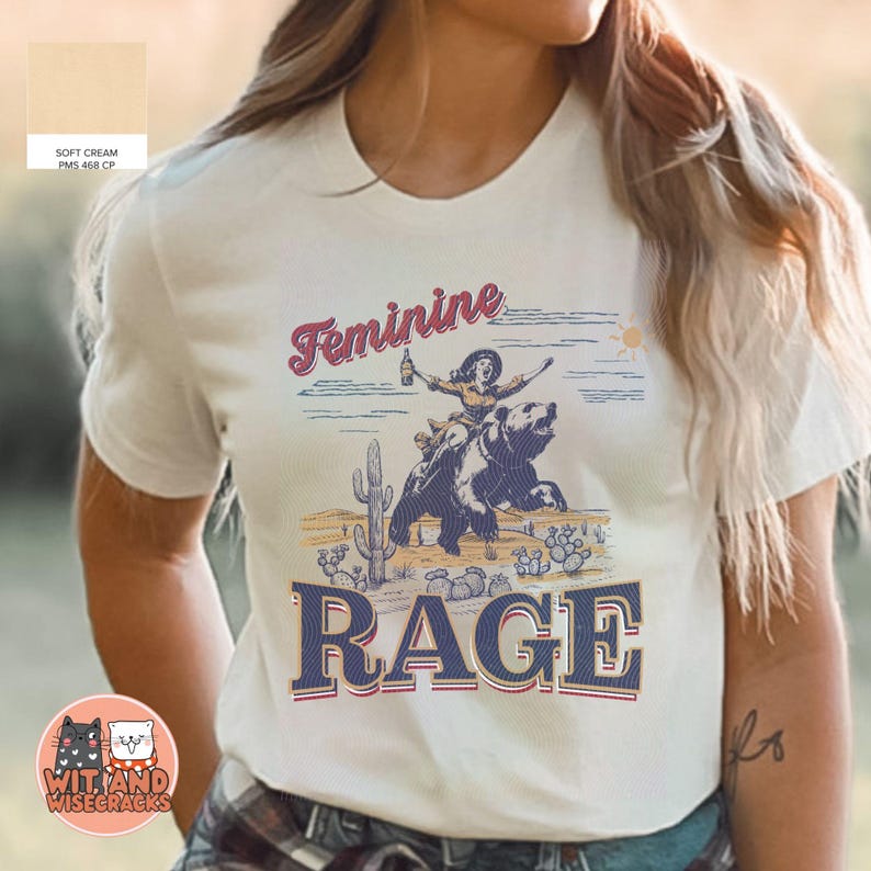 May include: A white t-shirt with a graphic design of a woman riding a bear. The graphic includes the text "Feminine Rage" and a cactus.