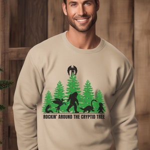 Puede incluir: Sudadera beige de cuello redondo con un gr&aacute;fico verde y negro de cr&iacute;ptidos y &aacute;rboles de Navidad. El texto "ROCKIN' AROUND THE CRYPTID TREE" est&aacute; impreso debajo. Un &aacute;rbol de Navidad es visible a la izquierda.