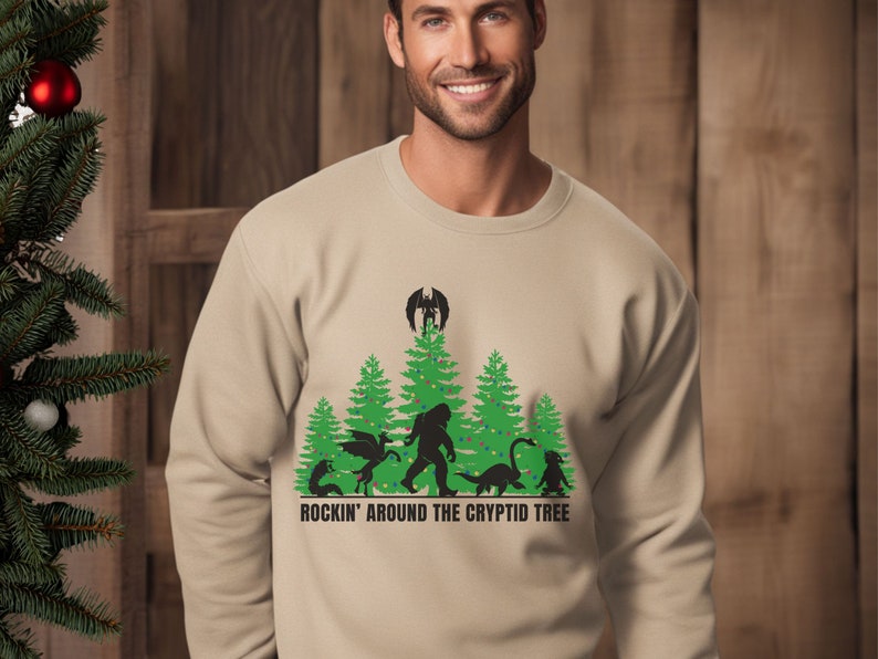 Puede incluir: Sudadera beige de cuello redondo con un gr&aacute;fico verde y negro de cr&iacute;ptidos y &aacute;rboles de Navidad. El texto "ROCKIN' AROUND THE CRYPTID TREE" est&aacute; impreso debajo. Un &aacute;rbol de Navidad es visible a la izquierda.