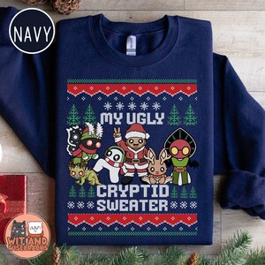May include: Navy blue sweatshirt with a festive design. The graphic reads "MY UGLY CRYPTID SWEATER" and features cartoon cryptids in Christmas attire. Red and white accents create a holiday sweater look.