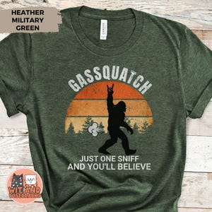 May include: Heather military green t-shirt with a graphic of a Bigfoot silhouette, the text "Gassquatch", and the phrase "Just one sniff and you'll believe". The design includes a sunset and trees.