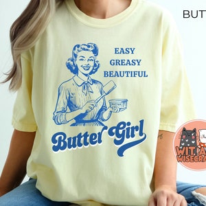 Pode incluir: Uma camiseta amarela com uma ilustra&ccedil;&atilde;o retr&ocirc; de uma mulher segurando uma barra de manteiga. O texto na camiseta diz "Easy Greasy Beautiful Butter Girl".