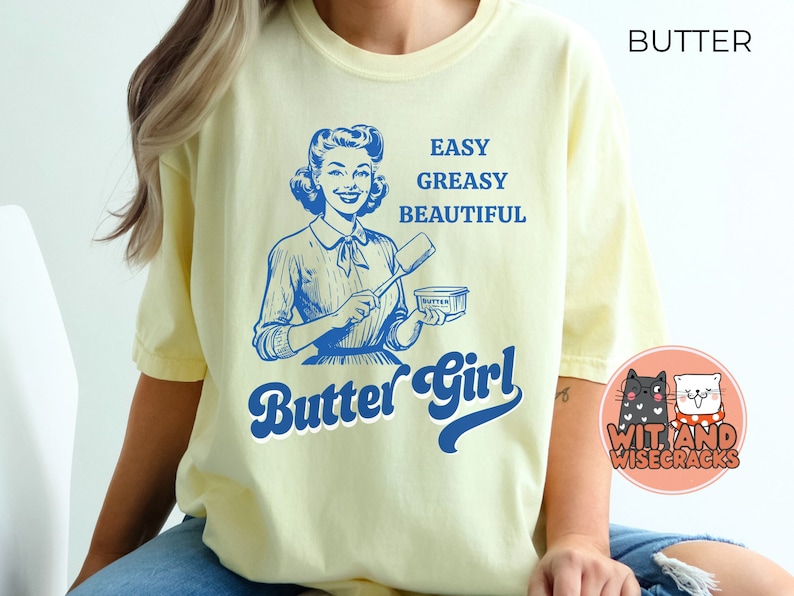 Pode incluir: Uma camiseta amarela com uma ilustra&ccedil;&atilde;o retr&ocirc; de uma mulher segurando uma barra de manteiga. O texto na camiseta diz "Easy Greasy Beautiful Butter Girl".