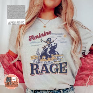 May include: A gray t-shirt with a graphic print of a woman riding a bear with the text "Feminine Rage" in a vintage style.
