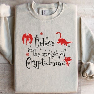 May include: A beige sweatshirt with the text "Believe in the magic of Cryptidmas" in black lettering. The text is surrounded by red illustrations of a monster, a plesiosaur, and a unicorn.