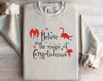May include: A beige sweatshirt with the text "Believe in the magic of Cryptidmas" in black lettering. The text is surrounded by red illustrations of a monster, a plesiosaur, and a unicorn.