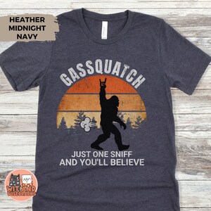 May include: Heather midnight navy t-shirt with a graphic of a Bigfoot silhouette giving a rock-on hand gesture in front of a sunset. Text reads "Gassquatch" and "Just one sniff and you'll believe."