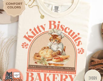 May include: A cream colored t-shirt with a vintage-style graphic of a cat wearing a chef's hat and baking biscuits. The graphic says "Kitty Biscuits" and "PAWS-ITIVELY DELIGHTFUL TREATS WE KNEAD LOVE INTO EVERY BATCH BAKERY".