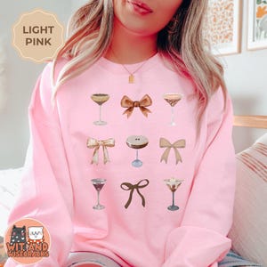 May include: A light pink sweatshirt with a graphic design of cocktails and bows. The design features six different cocktails and three bows in various shades of brown.