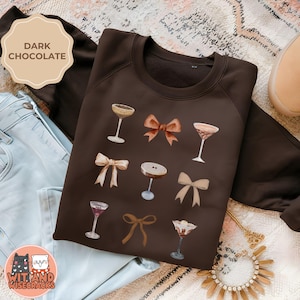 May include: A dark brown sweatshirt with a graphic design of cocktails and bows. The text "Wit and Wisecracks" is on the bottom left corner of the sweatshirt.