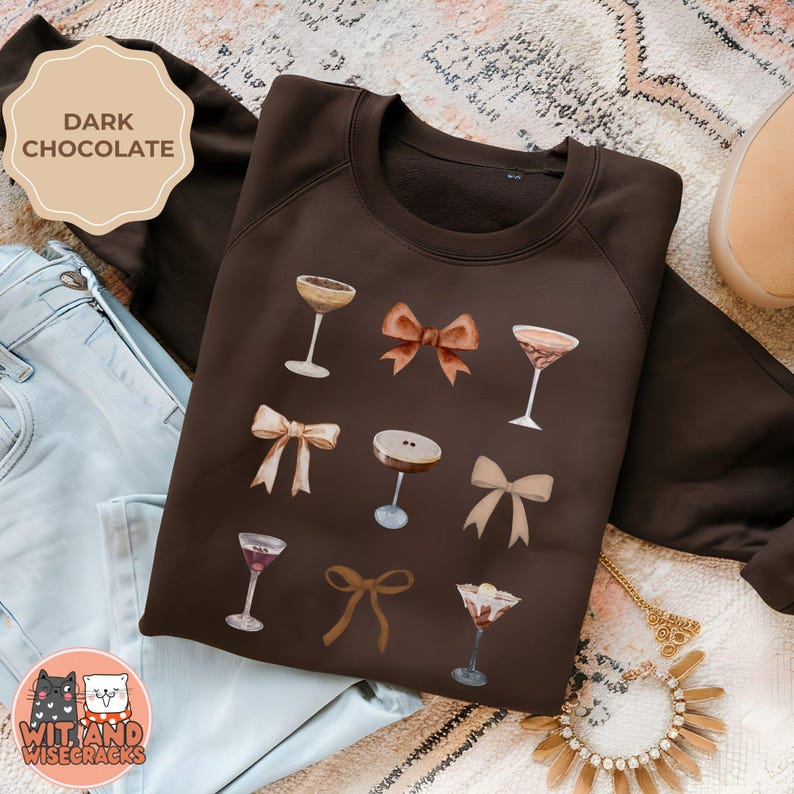 May include: A dark brown sweatshirt with a graphic design of cocktails and bows. The text "Wit and Wisecracks" is on the bottom left corner of the sweatshirt.