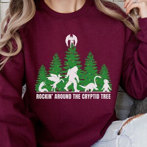 Puede incluir: Sudadera burdeos con un gr&aacute;fico de un &aacute;rbol de Navidad con cr&iacute;ptidos, incluyendo a Bigfoot y un drag&oacute;n. El texto "ROCKIN' AROUND THE CRYPTID TREE" est&aacute; impreso en blanco debajo.