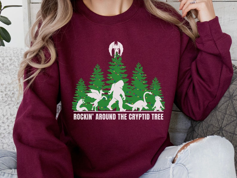 Puede incluir: Sudadera burdeos con un gr&aacute;fico de un &aacute;rbol de Navidad con cr&iacute;ptidos, incluyendo a Bigfoot y un drag&oacute;n. El texto "ROCKIN' AROUND THE CRYPTID TREE" est&aacute; impreso en blanco debajo.