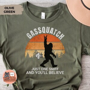 May include: Olive green t-shirt with a graphic of a Bigfoot silhouette, a sunset, and the text "Gassquatch." The design includes the phrase "Just one sniff and you'll believe."