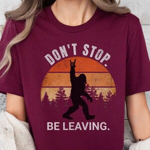 K&ouml;nnte beinhalten: Burgunderfarbenes T-Shirt mit einer Grafik im Vintage-Stil von Bigfoot mit einer Rock'n'Roll-Handgeste vor einem Sonnenuntergang. Der Text lautet "Don't Stop. Be Leaving."