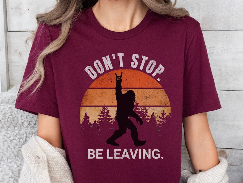 K&ouml;nnte beinhalten: Burgunderfarbenes T-Shirt mit einer Grafik im Vintage-Stil von Bigfoot mit einer Rock'n'Roll-Handgeste vor einem Sonnenuntergang. Der Text lautet "Don't Stop. Be Leaving."