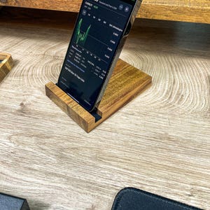 Könnte beinhalten: Ein Holz-Telefonständer mit einem schwarzen Smartphone, das darin liegt. Der Bildschirm des Telefons zeigt einen Aktienmarkt-Graphen mit Zahlen und Daten. Der Ständer steht auf einer hellen Holzoberfläche.