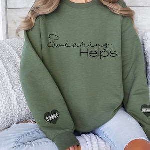 May include: A green sweatshirt with the text "Swearing Helps" printed on the front. The sweatshirt has a black heart with the word "CENSORED" printed on it on each sleeve.