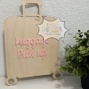 Può includere: Un ritaglio di legno di una valigia con la scritta "Luggage Pick Up" in lettere rosa. La valigia è su uno sfondo bianco con una pianta verde sullo sfondo.