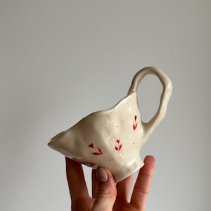May include: Cream-colored ceramic gravy boat with a handle and a flared rim. The boat is decorated with small red floral designs. The handle is a loop shape. The piece is held in a hand against a plain background.