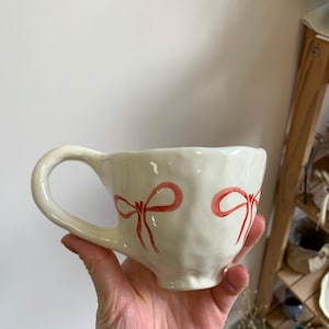 Puede incluir: Taza de cerámica blanca con superficie texturizada y lazos pintados de rojo. La taza tiene una forma redondeada y un asa.