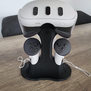 May include: A black stand with two white controllers and a white VR headset. The stand has a curved base and two arms that hold the controllers. The headset is resting on the top of the stand.