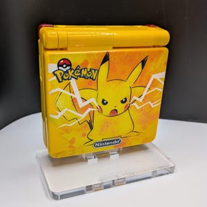 May include: Yellow Game Boy Advance SP with Pikachu and lightning bolt design. The handheld console has a red hinge and the Pokemon and Nintendo logos. It is displayed on a clear acrylic stand.