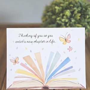 May include: A greeting card with the text "Thinking of you as you enter a new chapter in life." features an open book illustration with colorful pages and butterflies. The card is on a wooden surface.