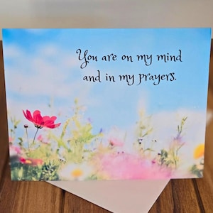 May include: A greeting card with a blue sky and white clouds background. The card features a pink flower and the text "You are on my mind and in my prayers."