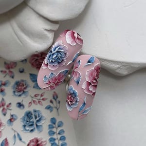May include: Two almond-shaped nails with a pale pink base, decorated with 3D floral designs. The flowers are blue, pink, and white, with blue and pink leaves. Floral nail art.