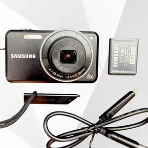 May include: A black Samsung digital camera with a 4.7-23.5mm 1:3.3-5.9 26mm zoom lens, a Samsung BP70A lithium-ion battery, a black wrist strap with a small plastic attachment, and a black USB charging cable with a multi-pin connector.