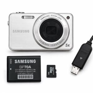 May include: A silver Samsung digital camera with a 4.7-23.5mm 5x zoom lens, accompanied by a black BP70A lithium-ion battery, a 2GB microSD memory card, and a black USB charging cable with a connector head featuring a triangular arrow icon.