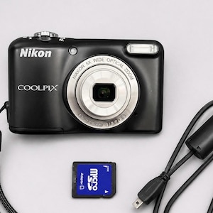 以下が含まれることがあります： シルバーのレンズリング、黒いリストストラップ、アダプター付きの青と黒のmicroSDカード、黒いUSB充電ケーブルを備えた黒いNikon Coolpixデジタルカメラ。カメラにはNikon、Coolpix、Nikkor 5x Wide Optical Zoomの文字が記載されています。