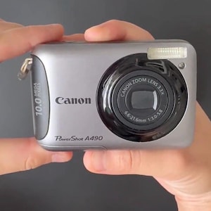 以下が含まれることがあります： 左側に10.0 Mega Pixelsのラベル、Canon Zoom Lens 3.3x 6.6-21.6mm 1:3.0-5.8と書かれた黒い円形のレンズハウジング、右上に小さな長方形のフラッシュを備えた、シルバーのCanon PowerShot A490デジタルカメラ。