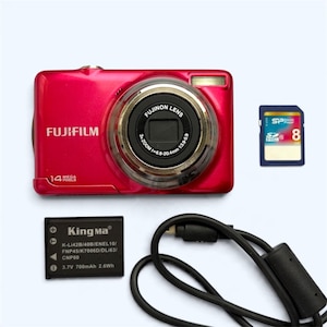 May include: A bright red Fujifilm digital camera with a 14 mega pixels label, a Fujinon lens, an 8GB Silicon Power SDHC memory card, a black KingMa 3.7V 700mAh 2.6Wh battery, and a black USB charging cable with a ferrite core filter.